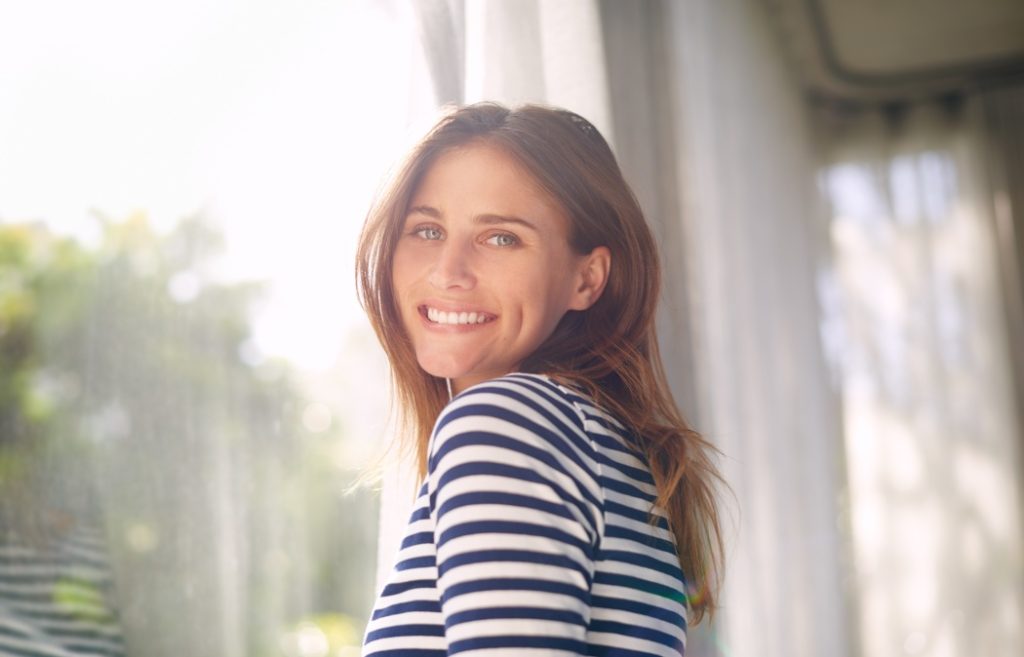 Woman, smile and portrait in house for relax and calm for holiday and cheerful on blurred background. Young person and happy with window for view of garden or backyard with sunlight in lounge